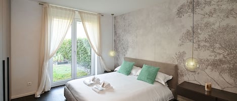 A modern bedroom with a large bed, two pillows, a nightstand, a wall-mounted lamp, and a view of greenery through the window.