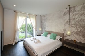 Ein modernes Schlafzimmer mit einem großen Bett, zwei Kissen, einem Nachttisch, einer Wandleuchte und Blick ins Grüne durch das Fenster.