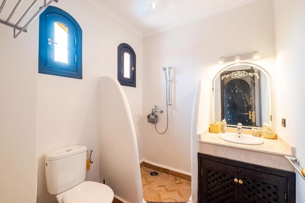 A bathroom with a white toilet, a shower, a sink with a mirror, and a small window with blue trim.