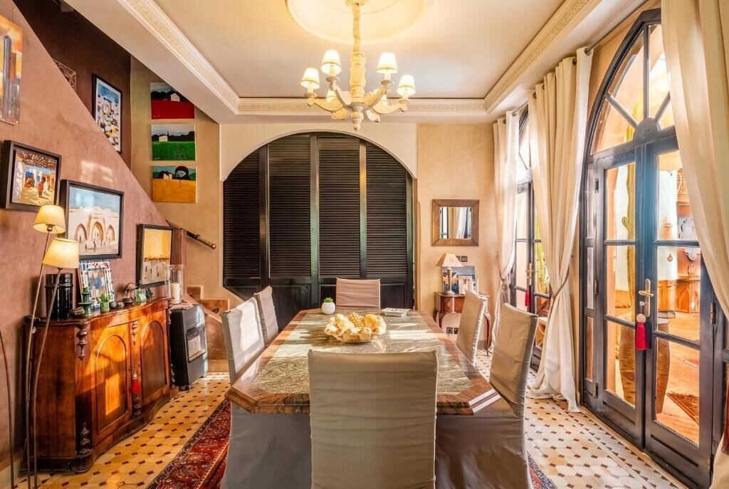 A dining area with a table set for four, a chandelier, and a view through a large window.