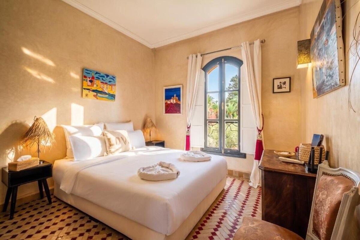 Ein Hotelzimmer mit einem großen Bett, einem Nachttisch, einem Schreibtisch, einem Sessel, einem Fenster mit Vorhängen und Bildern an den Wänden.