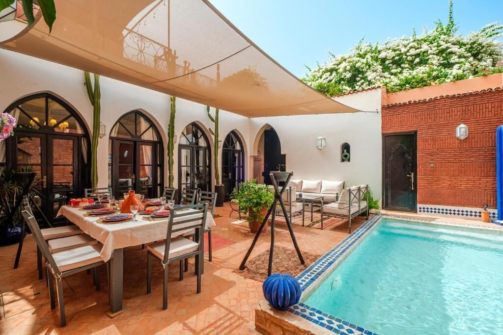 Uma área à beira da piscina com uma mesa de jantar posta para dois, cercada por janelas em arco e um alpendre coberto.