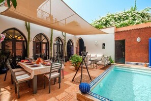 Un área junto a la piscina con una mesa de comedor para dos personas, rodeada de ventanales en arco y un porche cubierto.