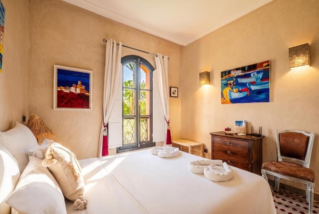 A hotel room with a bed, a desk, a chair, a window with curtains, and artwork on the walls.