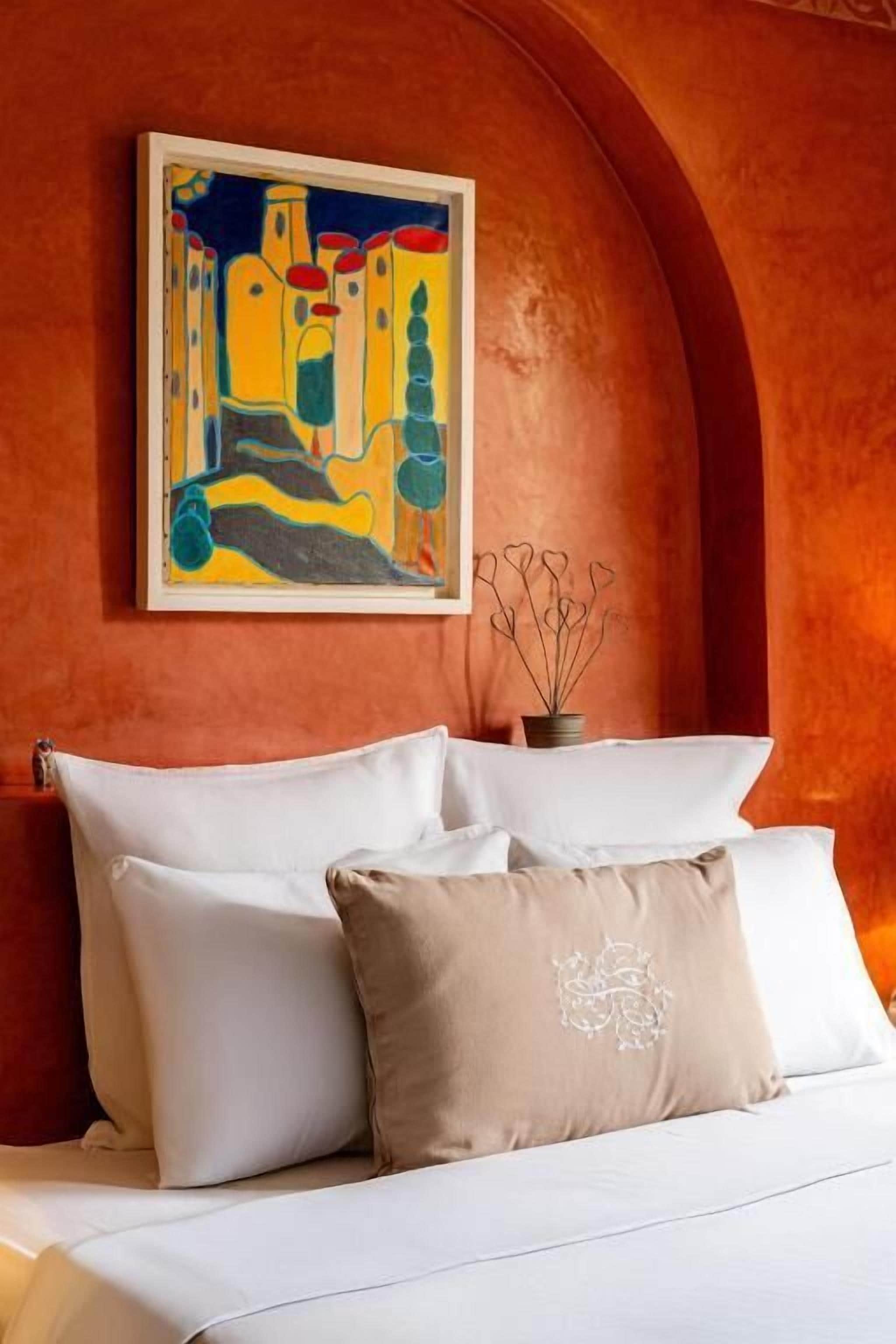 Una cama con sábanas blancas y almohada color beige, un cuadro enmarcado que muestra una escena de un pueblo colorido y un florero con ramas sobre una mesa.