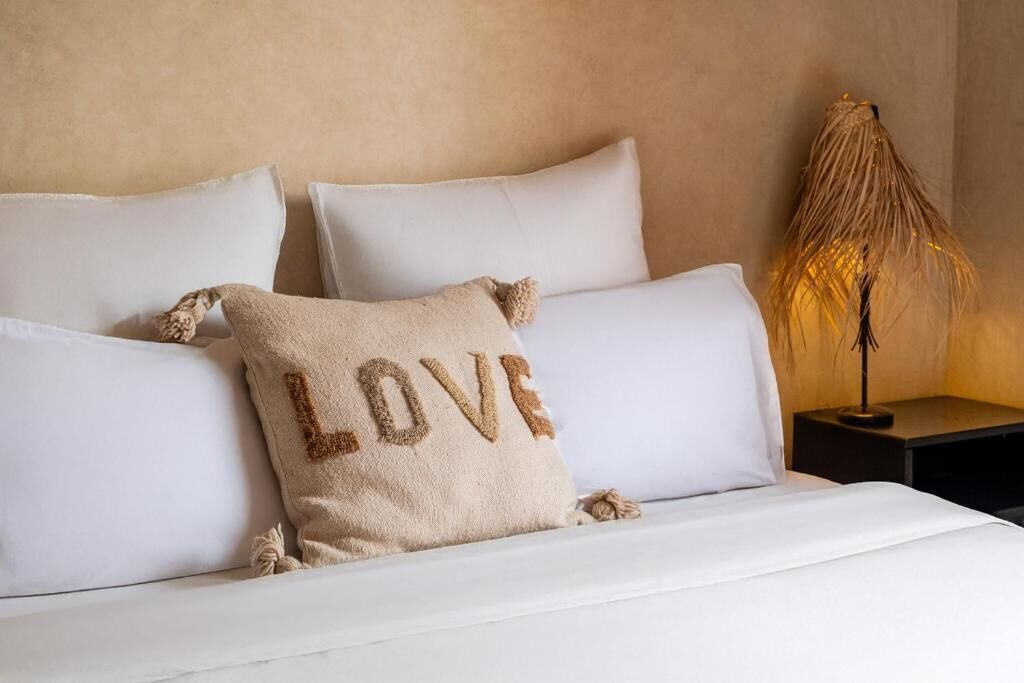 Una cama con sábanas blancas, un cojín beige con la palabra 'LOVE' bordada y una lámpara de noche.
