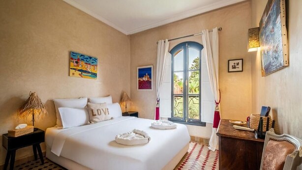 A hotel room with a large bed, a small table, a chair, a window with curtains, and artwork on the walls. - Riad Khiata (Marrakech)
