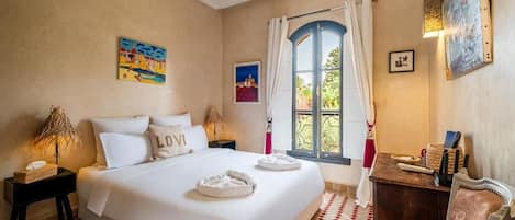 A hotel room with a large bed, a small table, a chair, a window with curtains, and artwork on the walls.