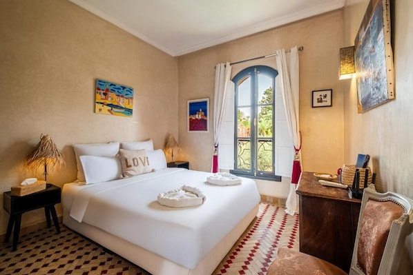Habitación de hotel con una cama grande, una mesita pequeña, una silla, una ventana con cortinas y obras de arte en las paredes.