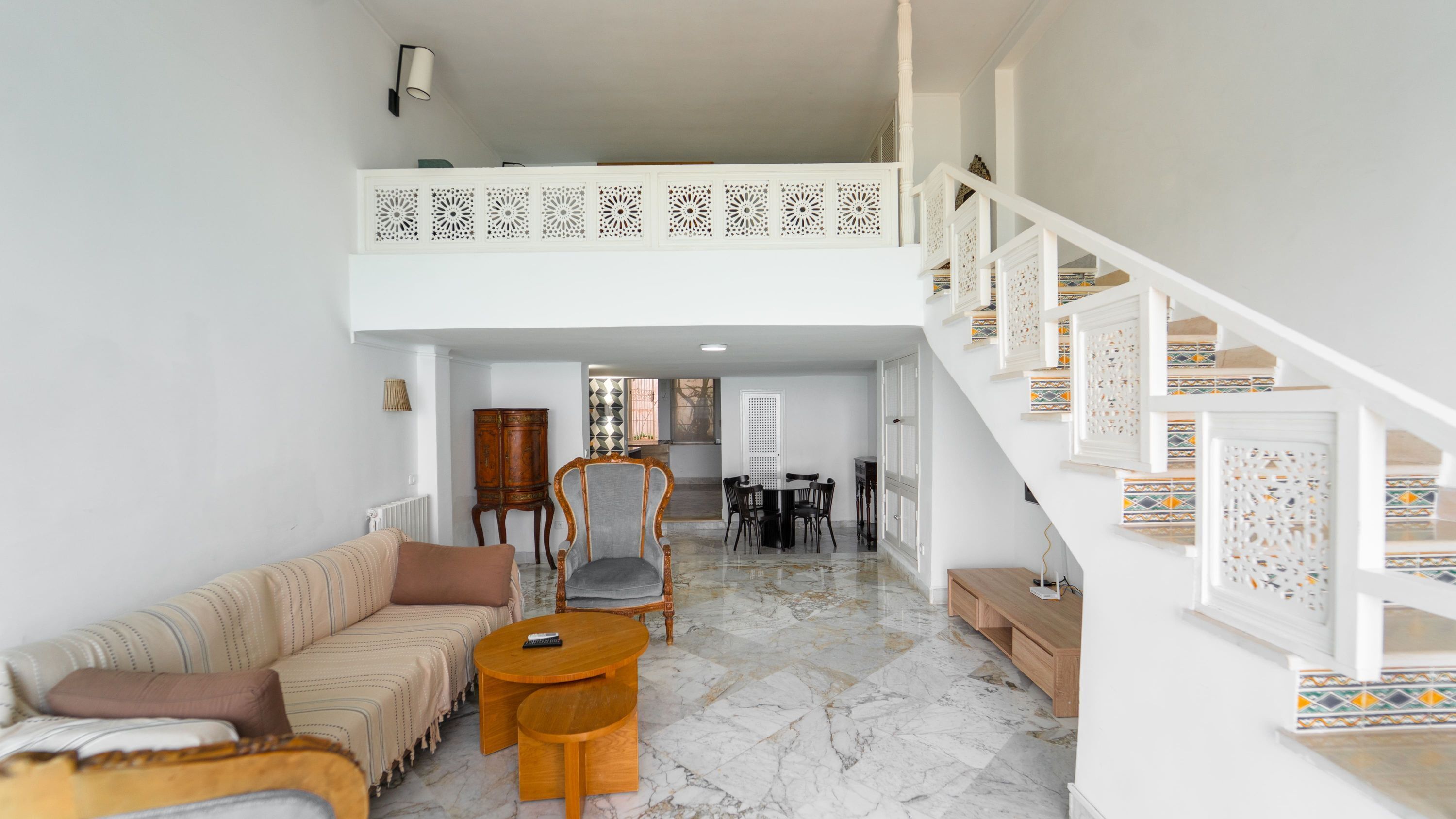 A spacious living area with a high ceiling, a staircase with decorative railings, a dining area, and a sofa set.