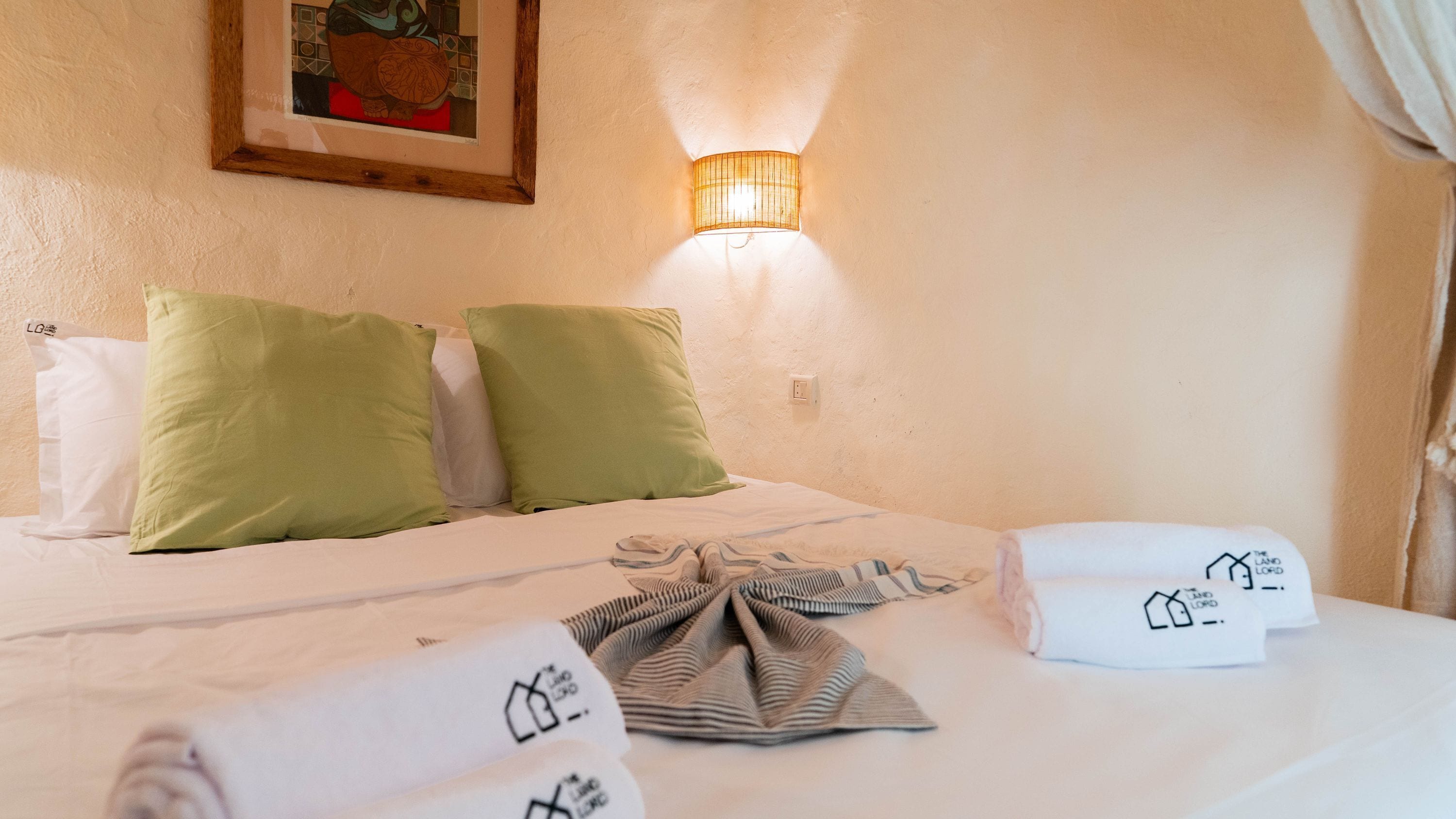 A hotel room with a bed, pillows, a wall-mounted light, and a painting.