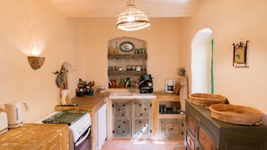 Cocina con estufa, lavaplatos y diversos utensilios. Se observa un lámpara colgante y una repisa con frascos y botellas.
