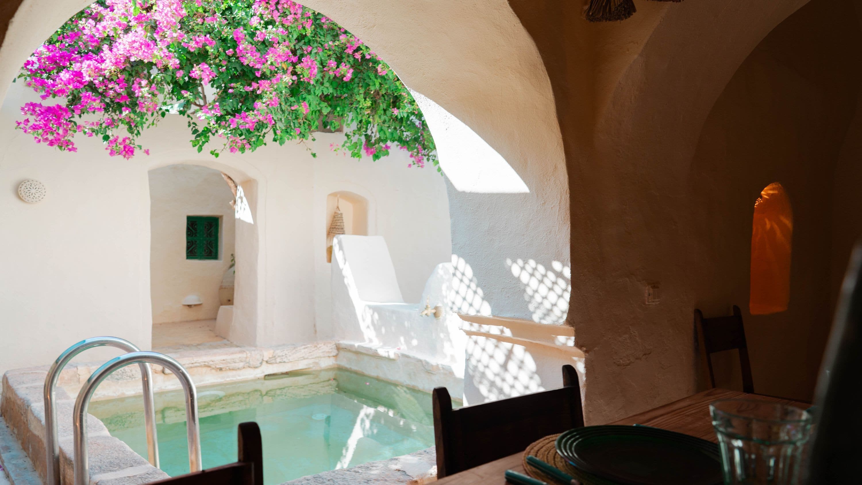 A sunlit pool area with a chair and a table, surrounded by white walls and arches.