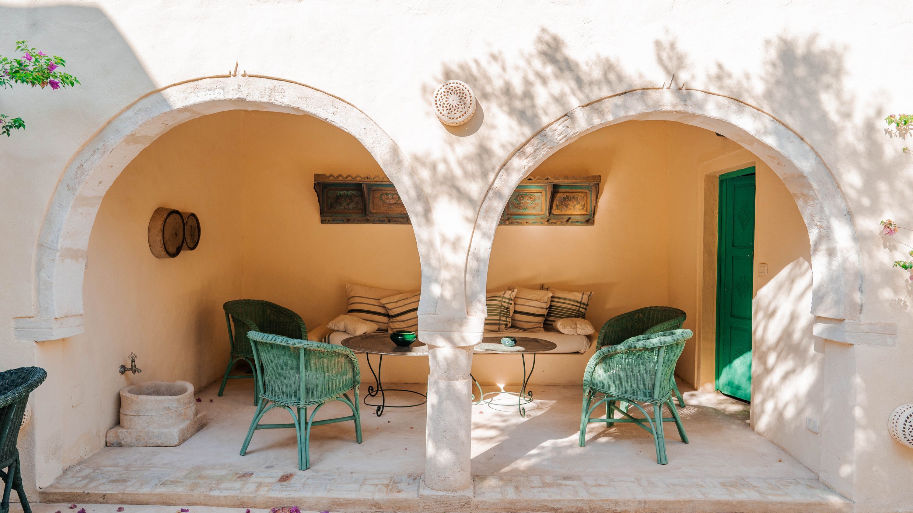 A cozy outdoor seating area with two green wicker chairs and a small table, under a white archway.