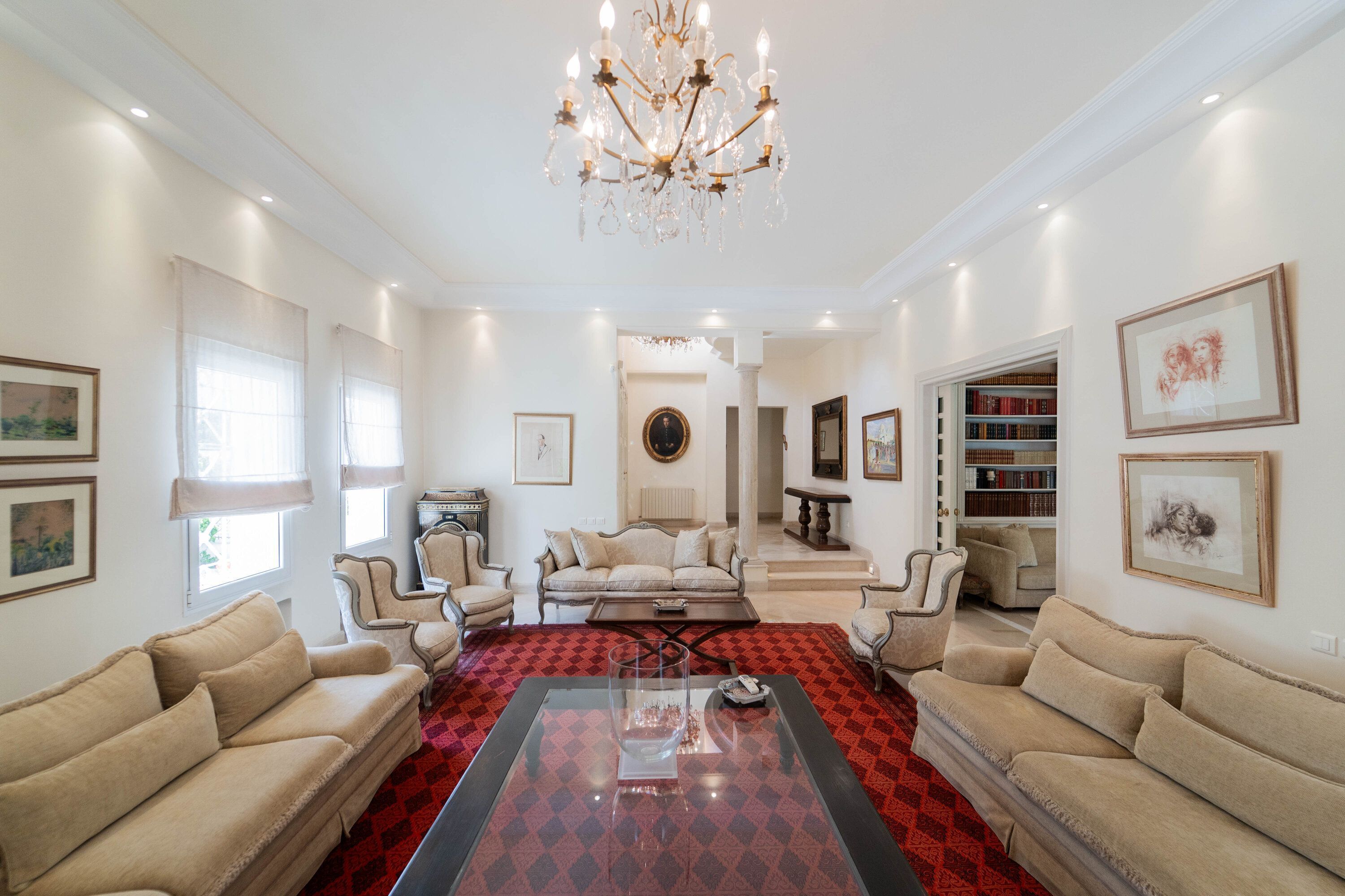 A spacious living room with a chandelier, multiple seating areas, and a red patterned rug.