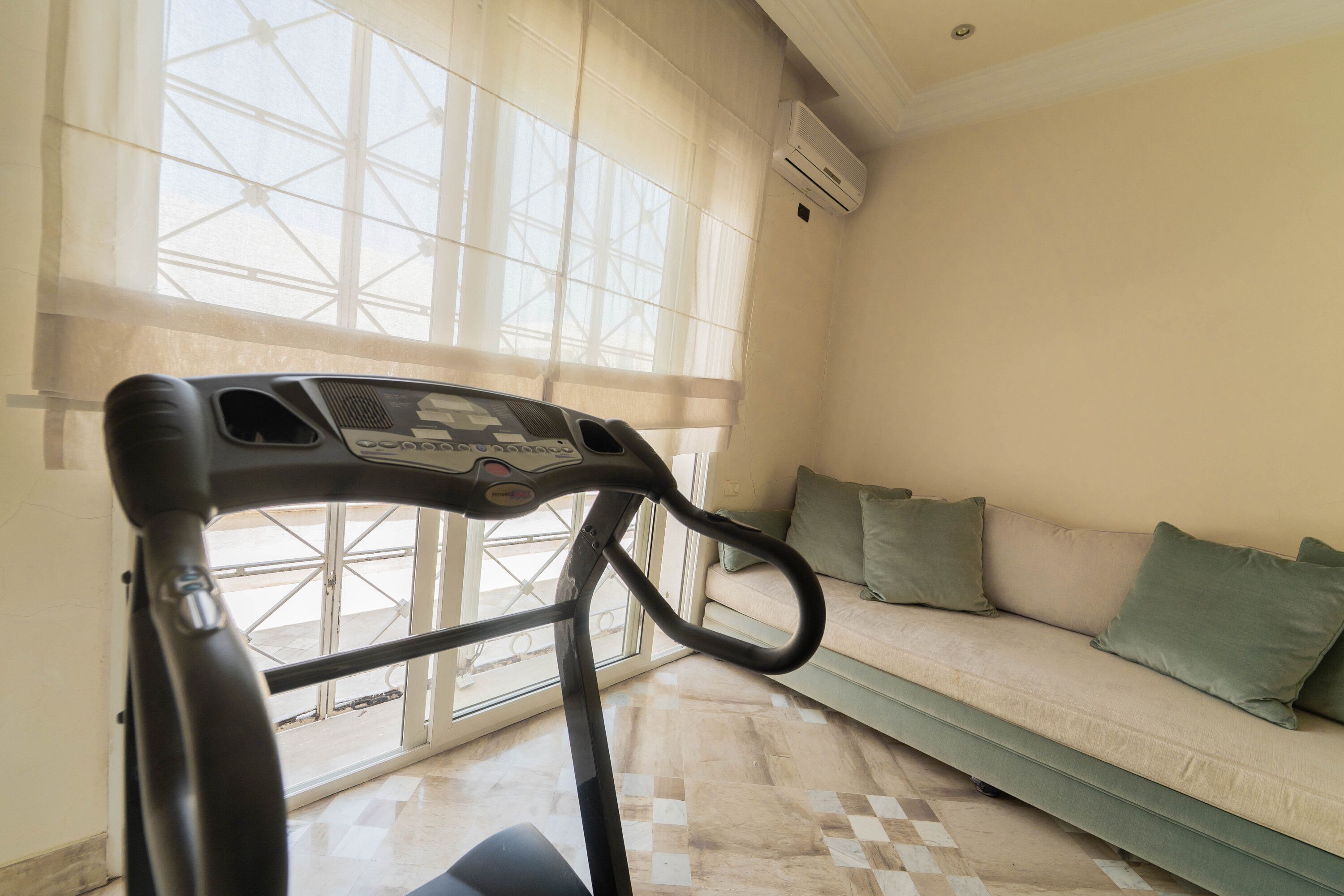 A stationary exercise bike in a room with a large window, a sofa, and a tiled floor.