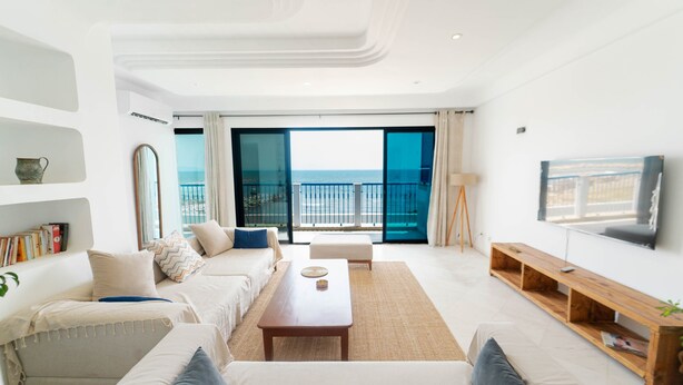 Ein modernes Wohnzimmer mit einer Sofagarnitur, einem Couchtisch, einem Fernseher und einem Balkon mit Meerblick.