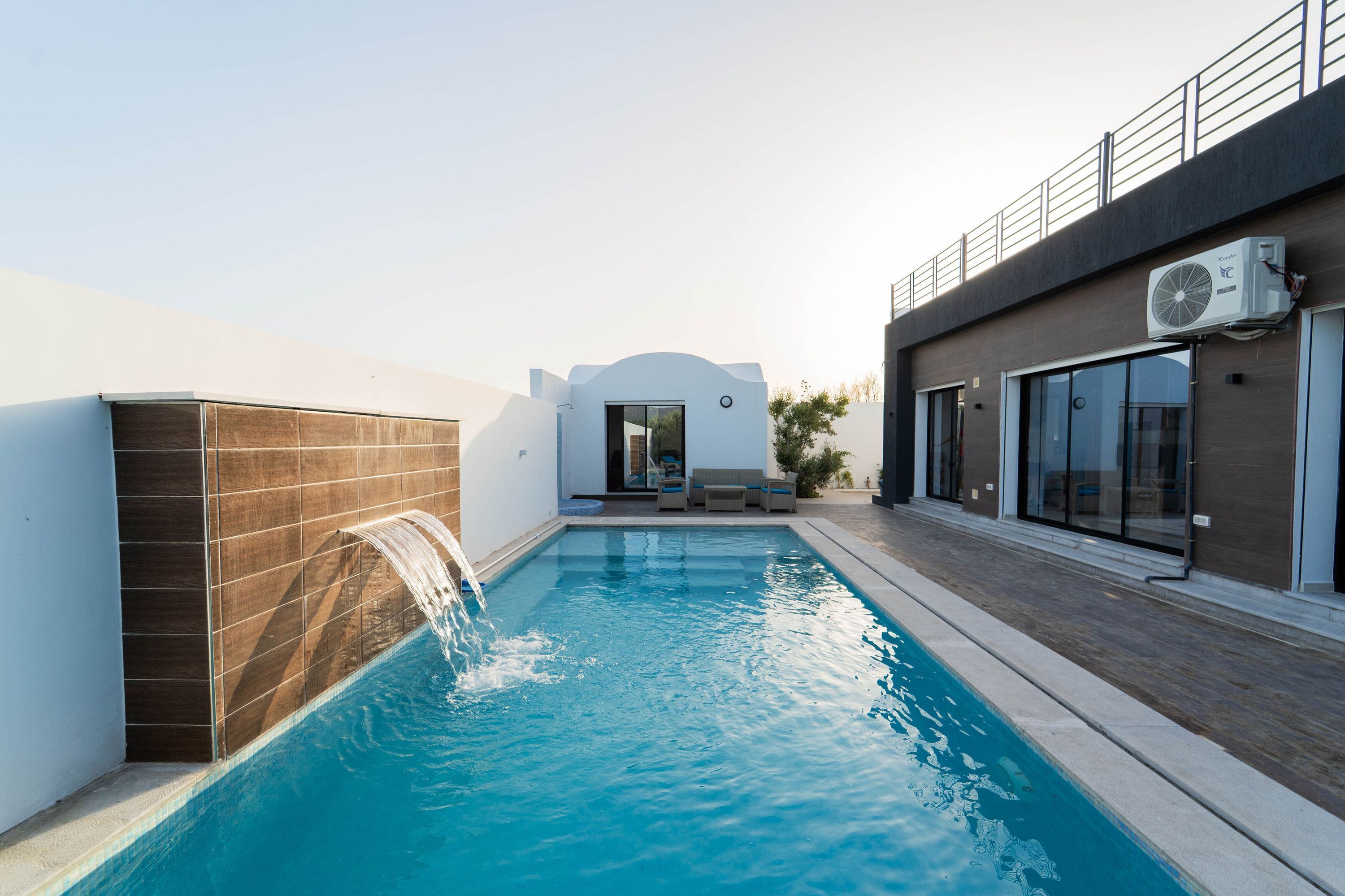 Una piscina con cascada, un área de estar cubierta y un edificio moderno con ventanales grandes.