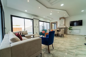 A spacious living area with a sofa, chairs, a dining table, and a flat-screen TV mounted on the wall. - 3-bedroom Villa With Private Pool Studio (Hammamet)