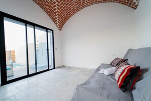 A modern living room with a grey sofa, a patterned throw blanket, and a view of a swimming pool through a glass door. - 3-bedroom Villa With Private Pool Studio (Hammamet)
