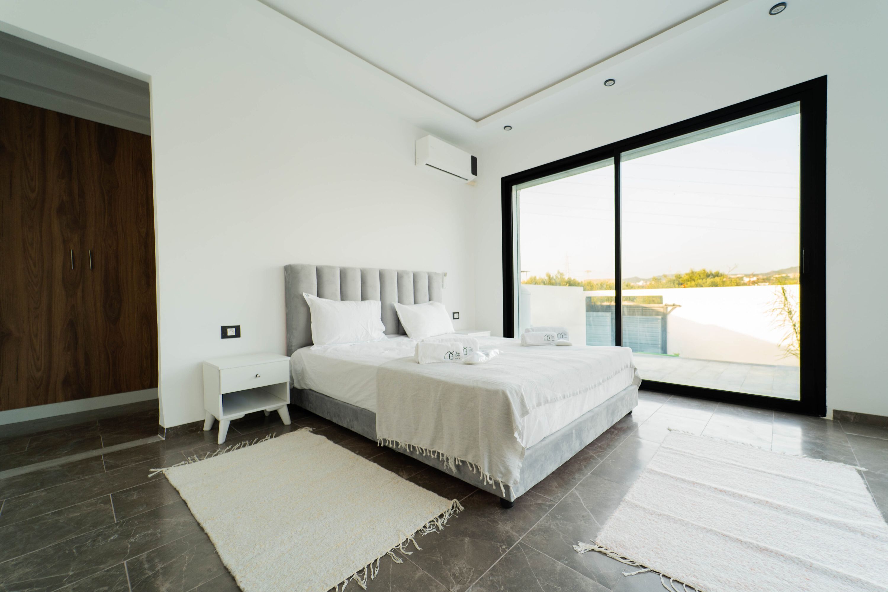 Un dormitorio moderno con una cama grande, una mesita de noche, un armario y vistas a través de un amplio ventanal.