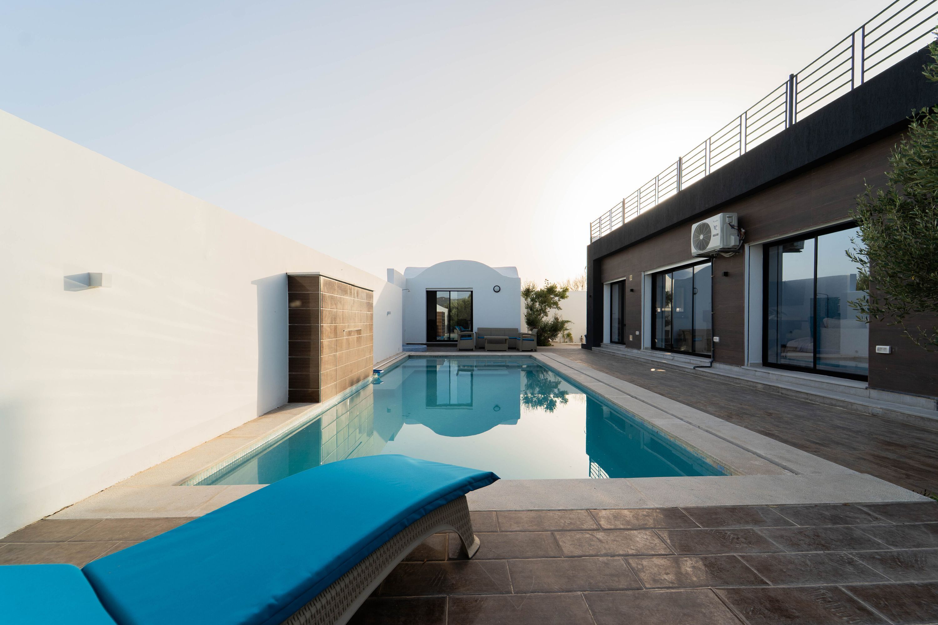 Una piscina esterna moderna con una sedia a sdraio, un muro in mattoni e un edificio con ampie finestre e un balcone.