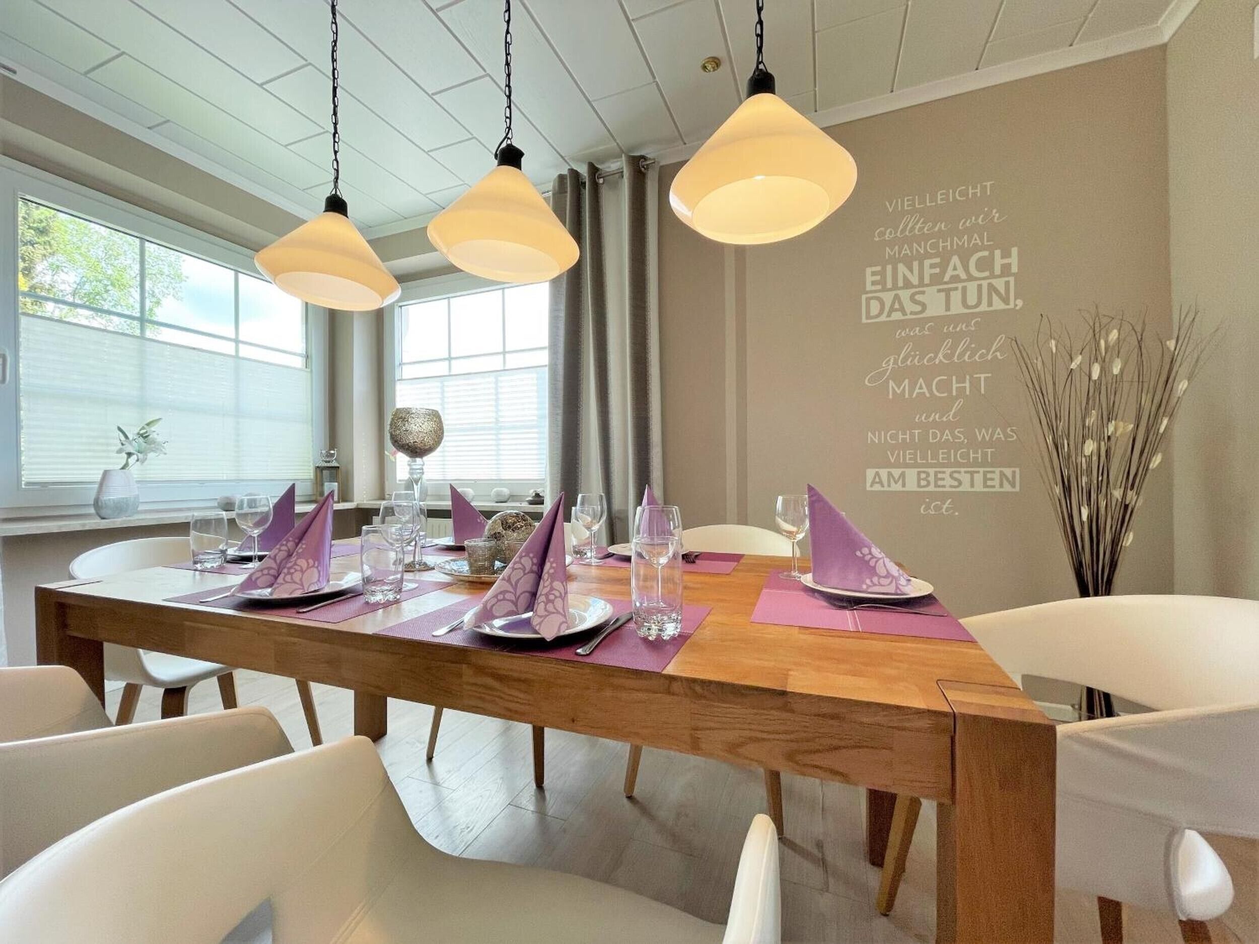 A dining area with a wooden table set for four, white chairs, and pendant lighting.