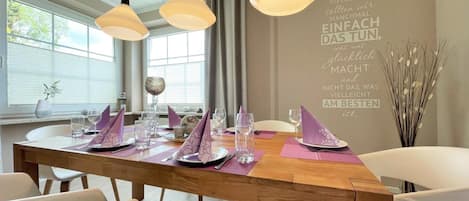 A dining area with a wooden table set for four, white chairs, and pendant lighting.