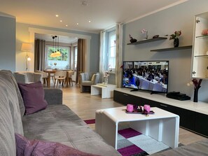 A modern living room with a sofa, coffee table, and a television displaying a conference. - SweetDreams Holiday Home – Terrace, Garden, Wi-Fi, Dogs Welcome – Private Guests Only (Bad Sachsa)