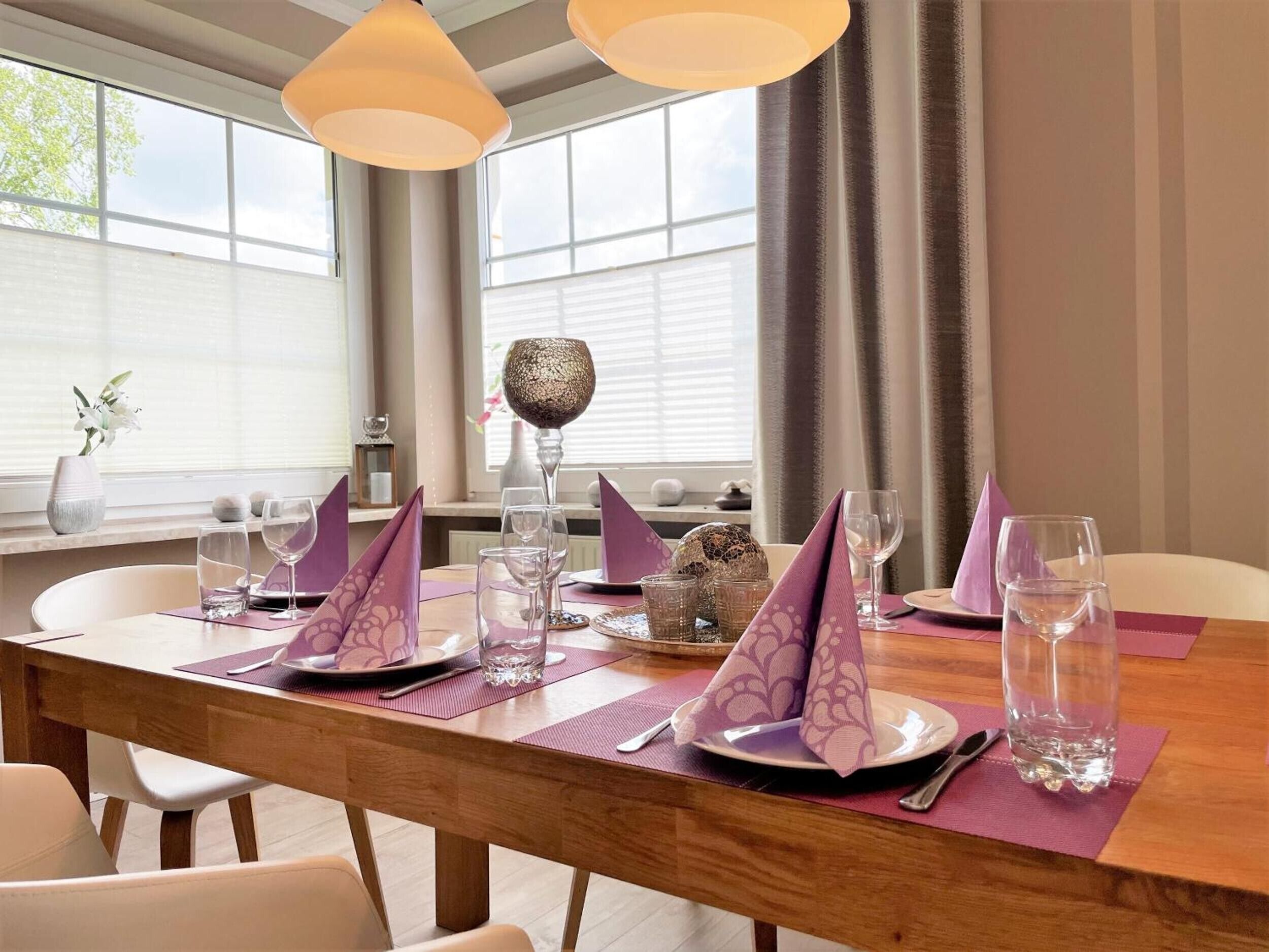 Een eettafel gedekt met paarse servetten, glazen en borden, met een vaas en decoratieve objecten op de vensterbank.
