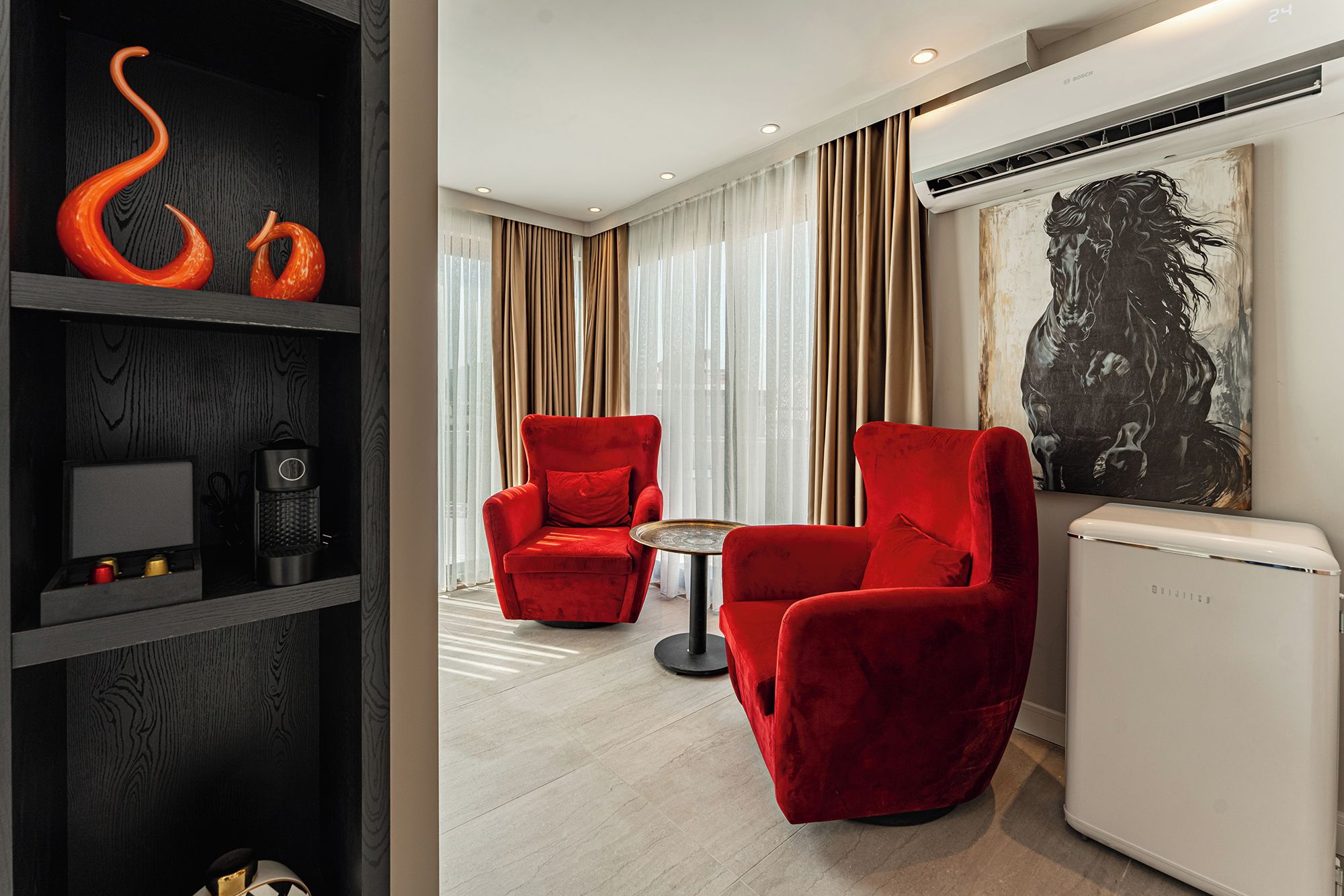 Une chambre d’hôtel moderne avec un fauteuil rouge, une petite table ronde et un grand tableau abstrait accroché au mur.
