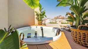 Un jacuzzi en una terraza de madera con plantas en macetas y vistas a edificios al fondo.