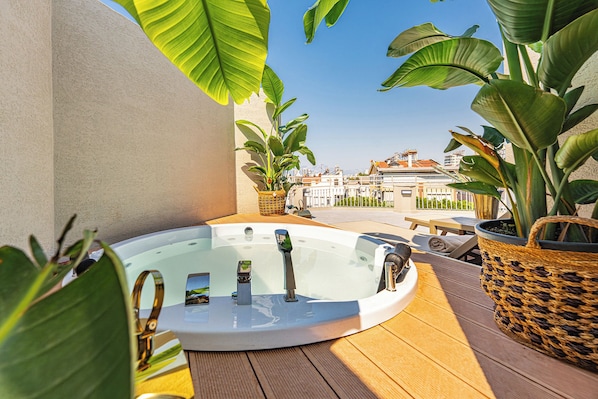 A hot tub on a wooden deck with potted plants and a view of buildings in the background.