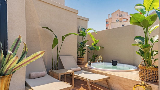 Un patio exterior iluminado por el sol con un jacuzzi, sillones de descanso y diversas plantas en macetas.