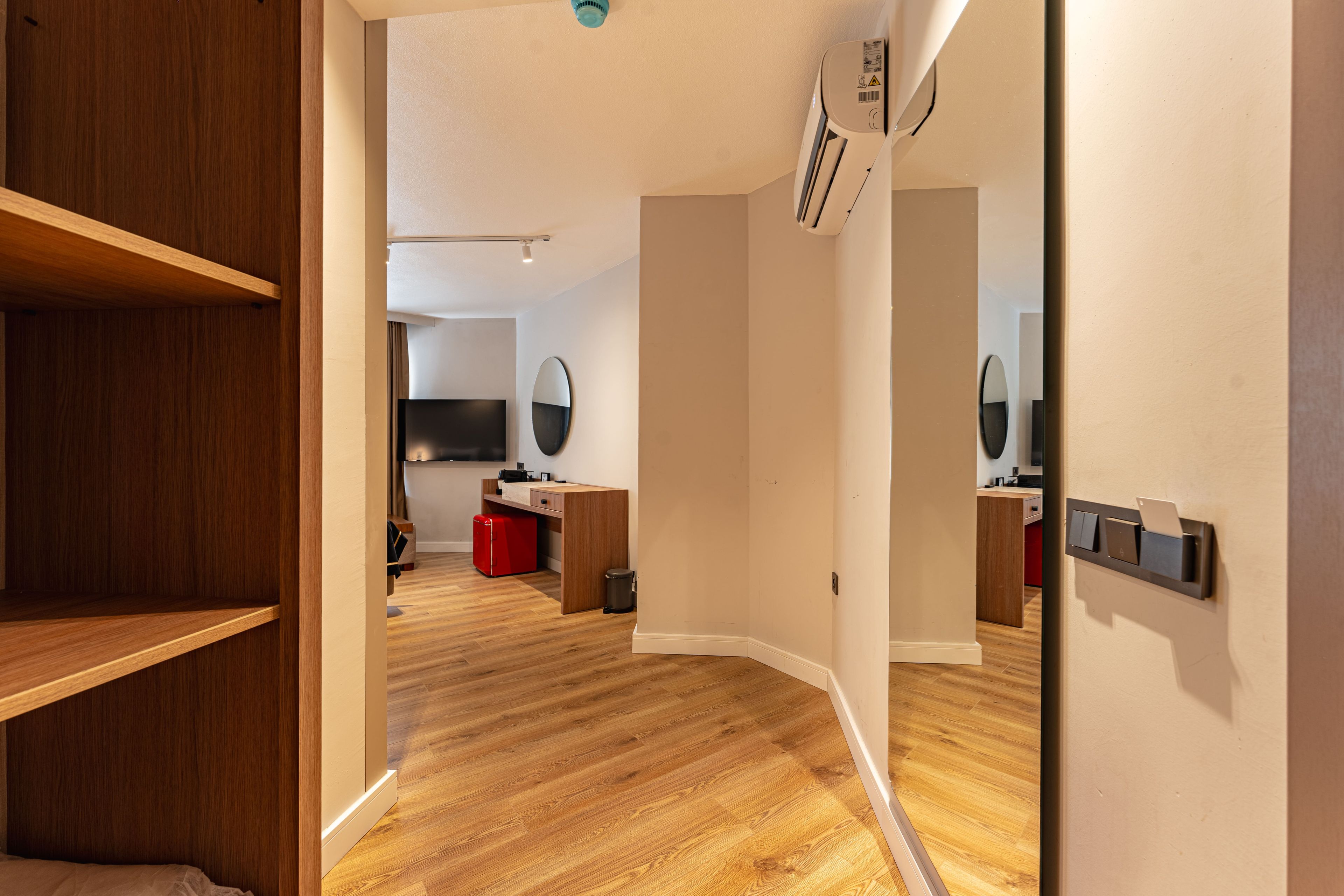A modern room with a wooden floor, a built-in wardrobe, a desk with a chair, a small trash bin, and a mirror on the wall.
