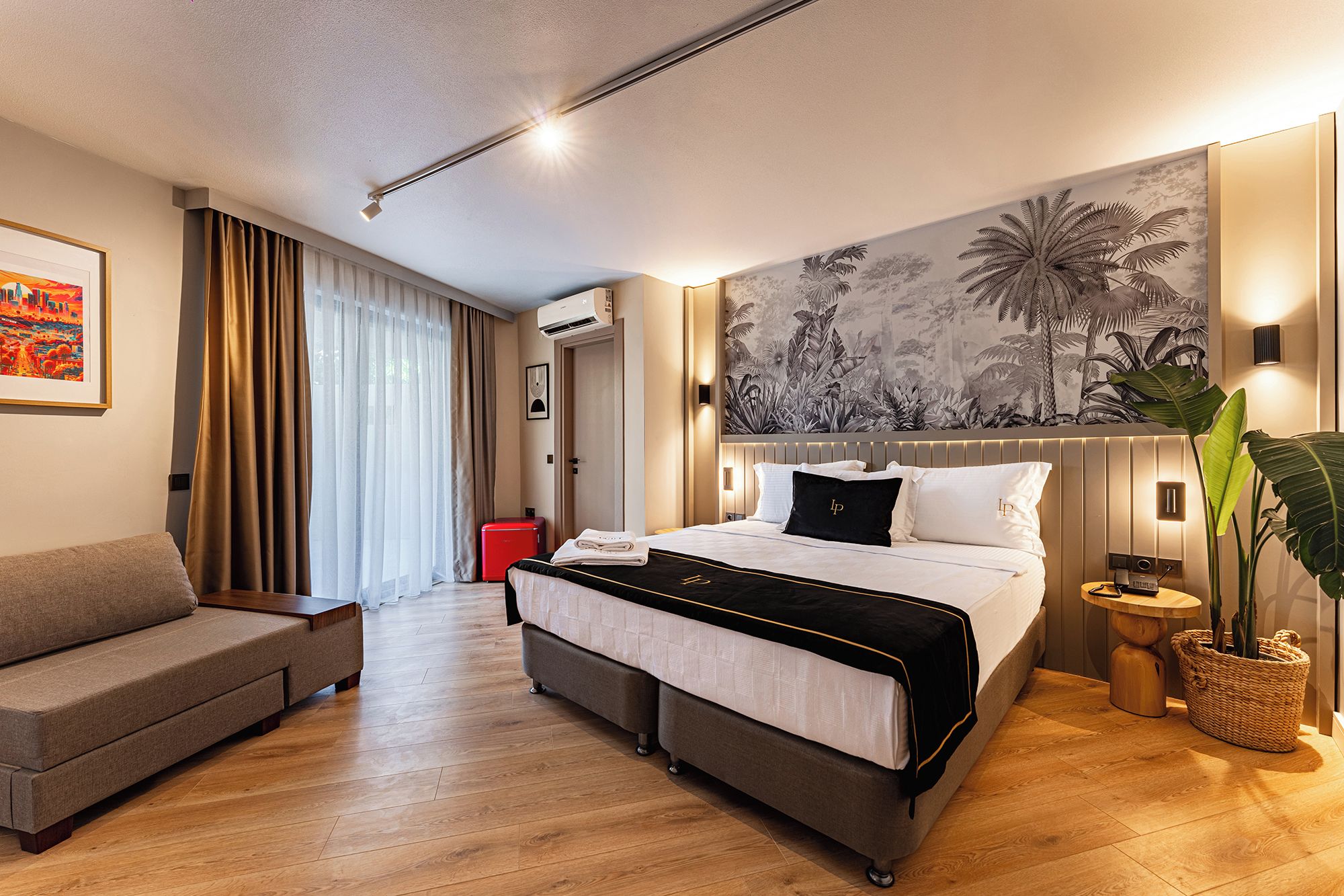 Habitación de hotel con cama, sofá, ventana grande con cortinas, aire acondicionado de pared, una obra de arte en la pared y una planta en maceta.