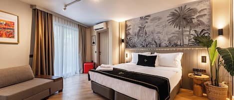 Habitación de hotel con cama, sofá, ventana grande con cortinas, aire acondicionado de pared, una obra de arte en la pared y una planta en maceta.