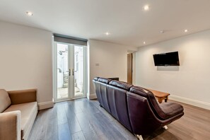 Ein modernes Wohnzimmer mit einer Couch, einem Liegesessel, einem an der Wand befestigten Fernseher und einer Schiebetür zu einem Außenbereich.