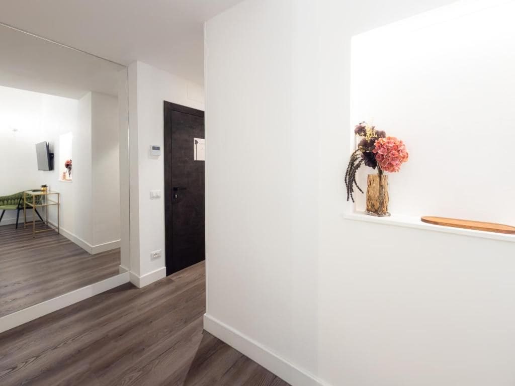 A hallway with a wooden floor, a door, and a wall-mounted shelf with a vase of flowers.