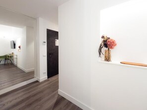 A hallway with a wooden floor, a door, and a wall-mounted shelf with a vase of flowers.