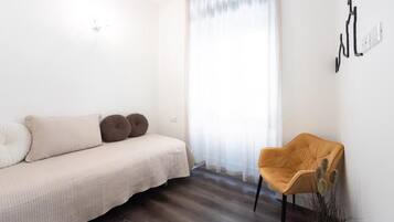 A minimalist room with a sofa, a chair, a window with sheer curtains, and a wall-mounted light switch.