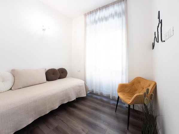 A minimalist room with a sofa, a chair, a window with sheer curtains, and a wall-mounted light switch.