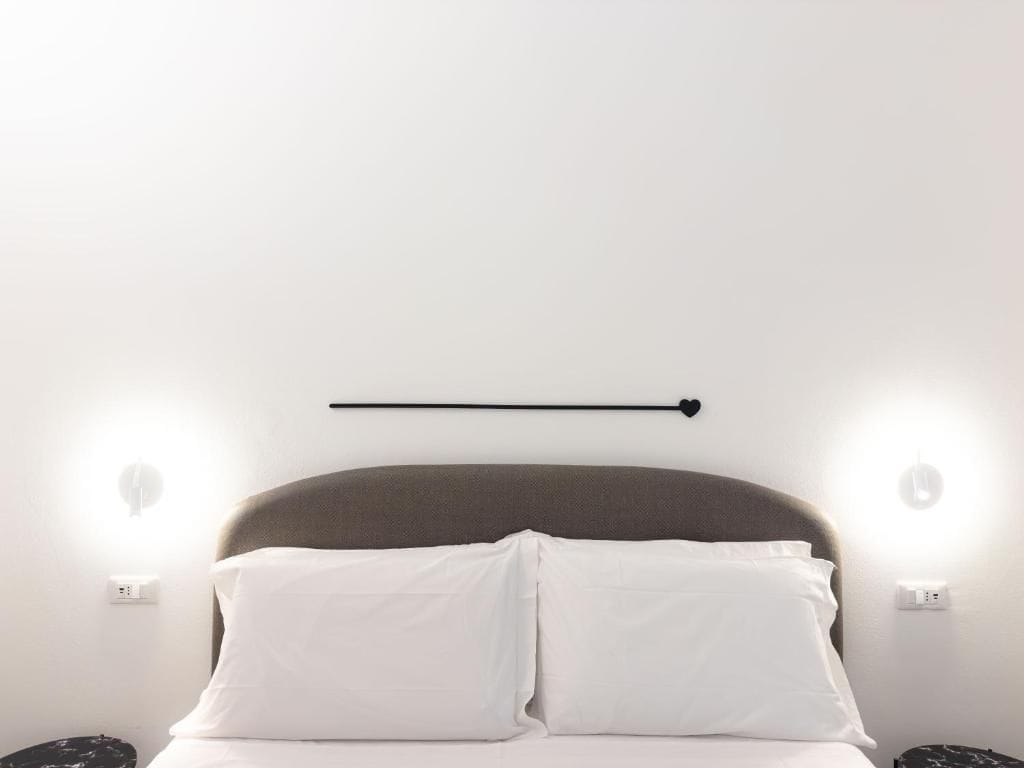 Een bed met witte beddengoed en een hoofdeinde, twee bedlampjes en een wandlamp.