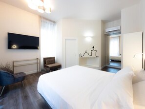 A hotel room with a bed, a flat-screen TV mounted on the wall, a seating area with a chair, and a wall-mounted artwork.
