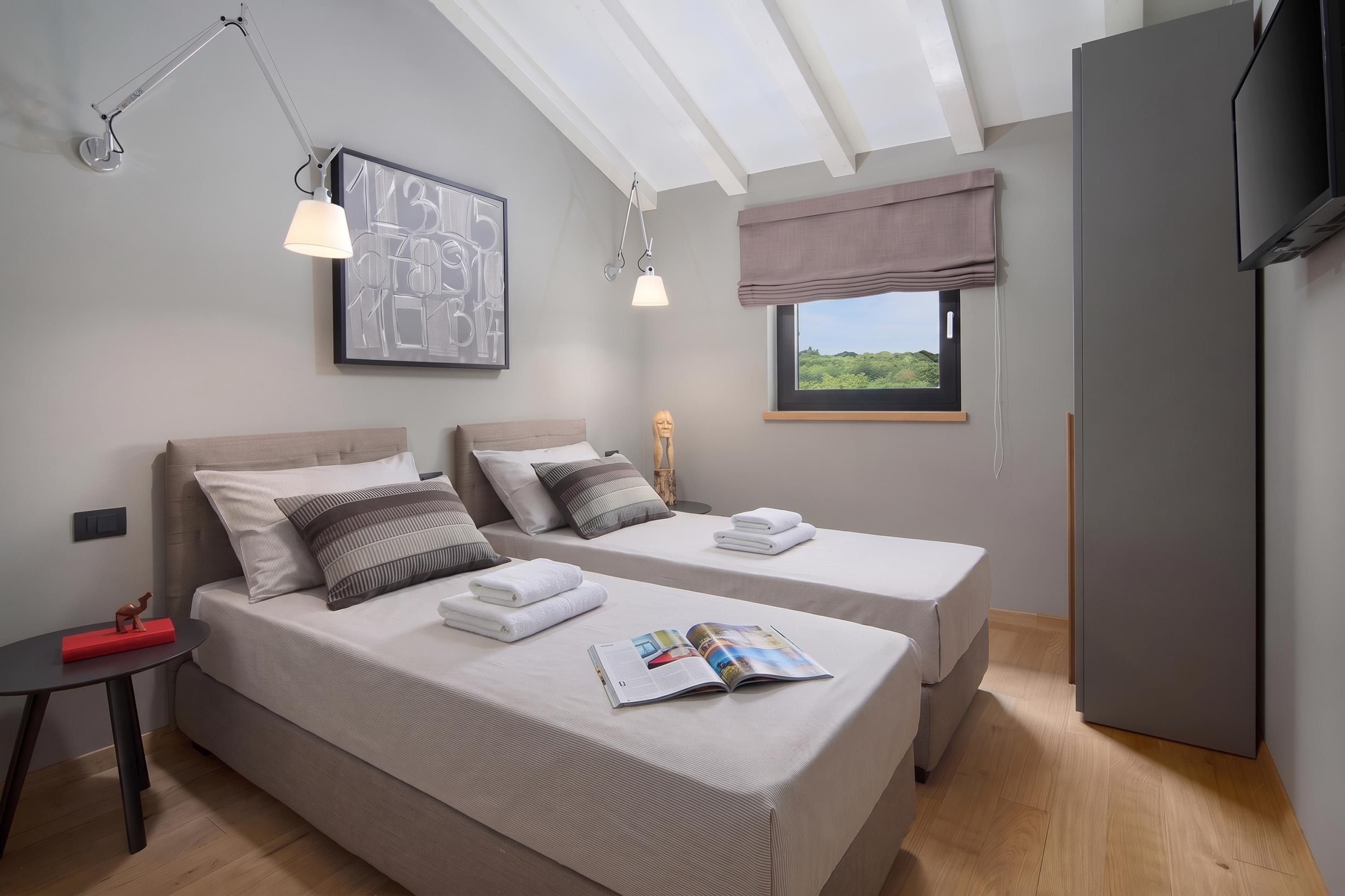 A modern bedroom with two beds, a window with a view, a flat-screen TV, and a ceiling light fixture.