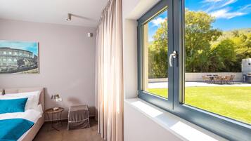 Habitación de hotel con cama, una mesita de noche con lámpara, una silla, una ventana con cortinas y vista a un área con césped.