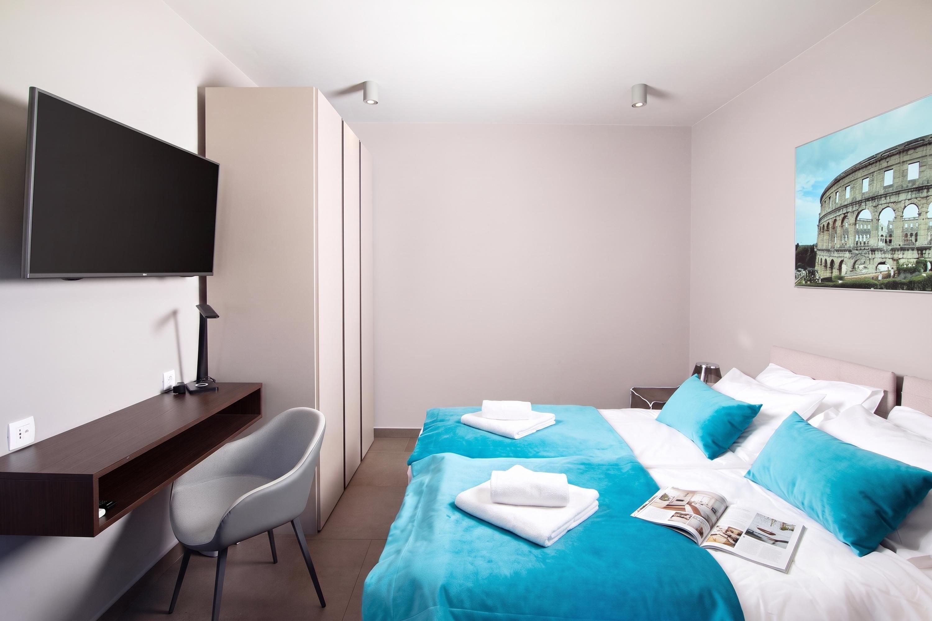 A hotel room with a bed, a desk, a chair, a television, and a painting of a historical building.