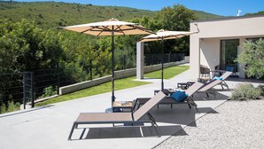 An outdoor patio with sun loungers and umbrellas, a modern building, and a hillside in the background. - Villa Awesome Lodge (Rasa)