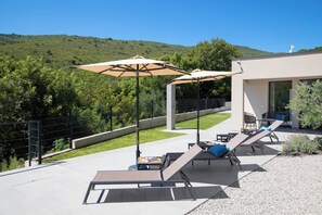 An outdoor patio with sun loungers and umbrellas, a modern building, and a hillside in the background. - Villa Awesome Lodge (Rasa)