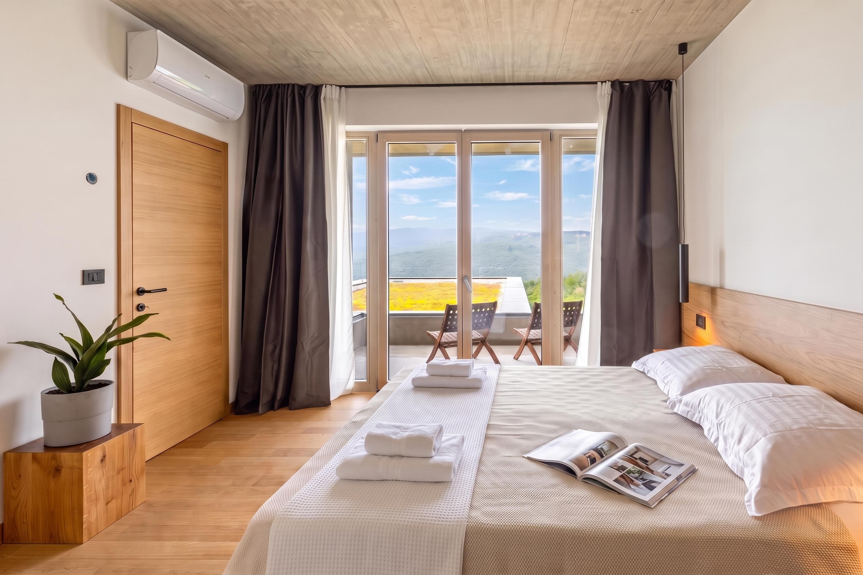 A bedroom with a bed, a potted plant, a window with curtains, and a view of a balcony.